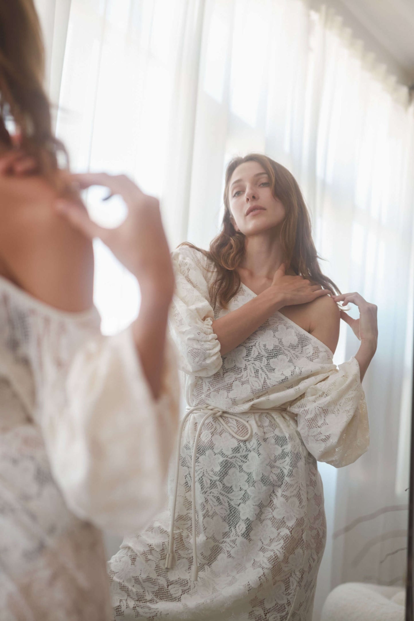 Woman in a lightweight ecru lace boho maxi dress with balloon sleeves, admiring herself in a mirror.