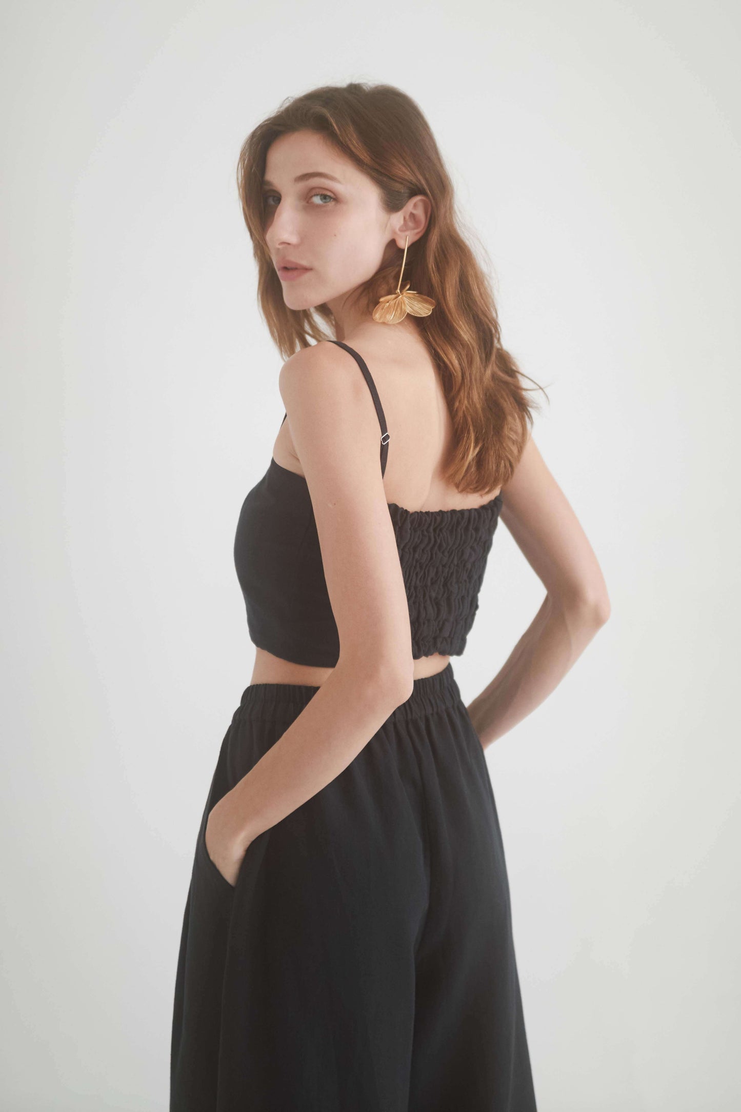 Model wearing a black eco-friendly crop top with adjustable straps and shirring detail, styled with wide trousers.