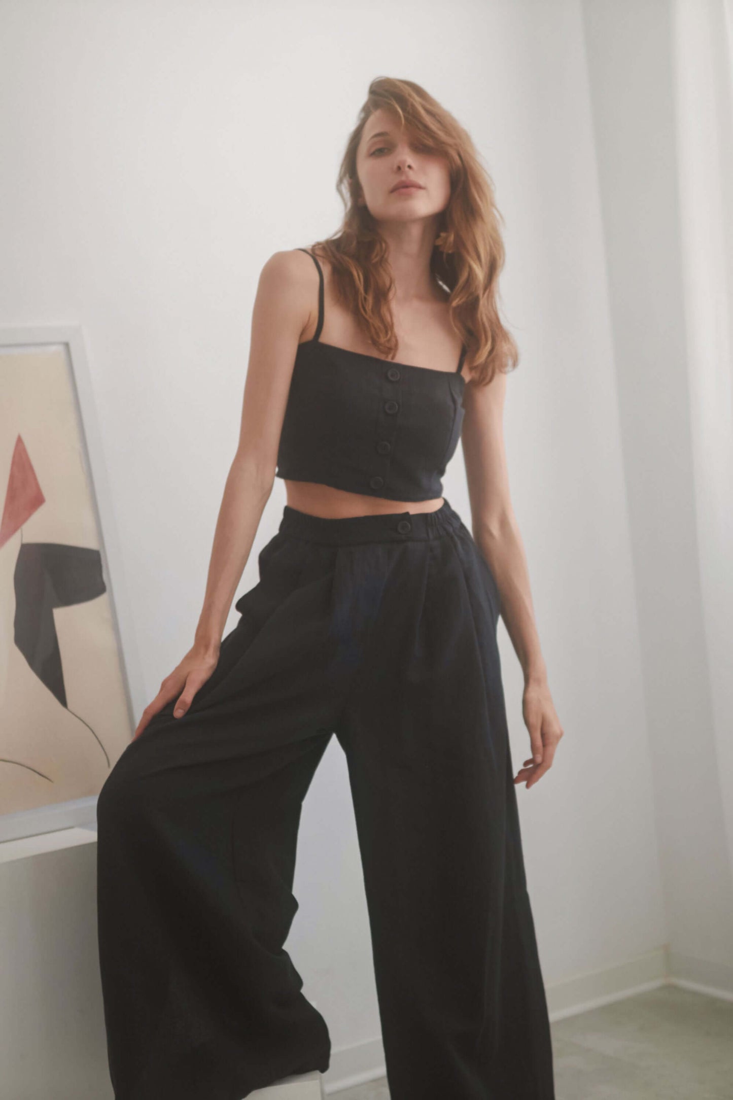 Model wearing a black crop top with adjustable straps and eco buttons, paired with wide trousers in a minimalistic setting.
