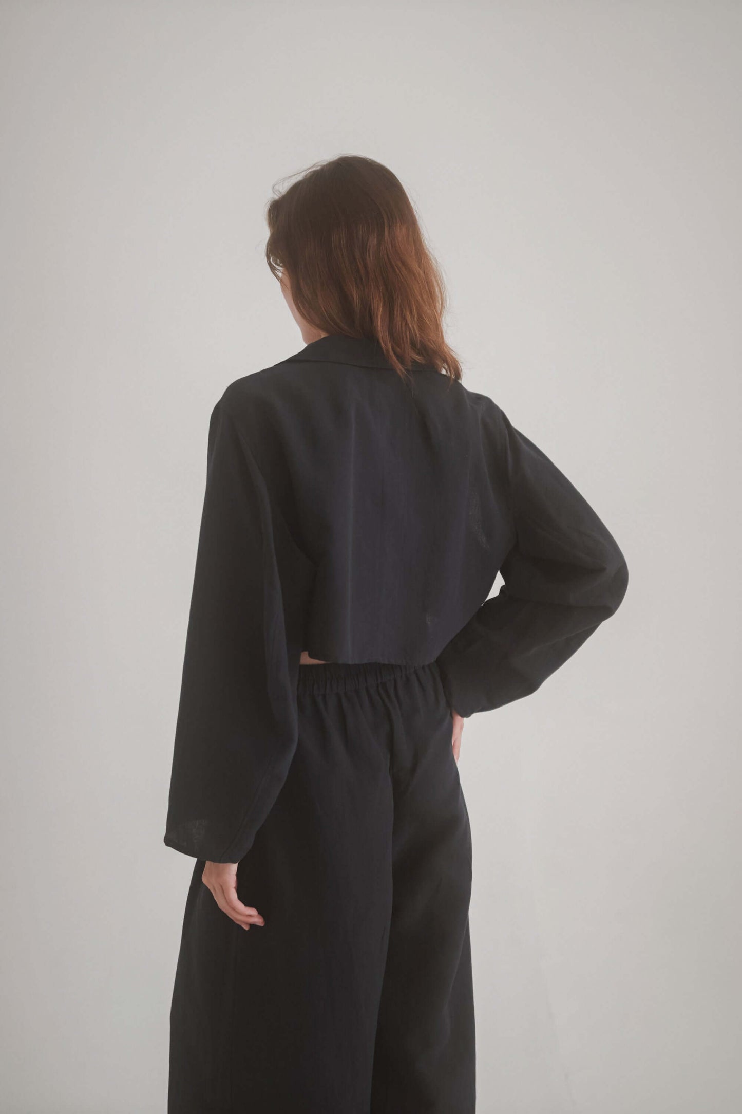 Back view of a woman wearing a black cropped jacket with wide trousers, showcasing the stylish fit and armhole cutouts.