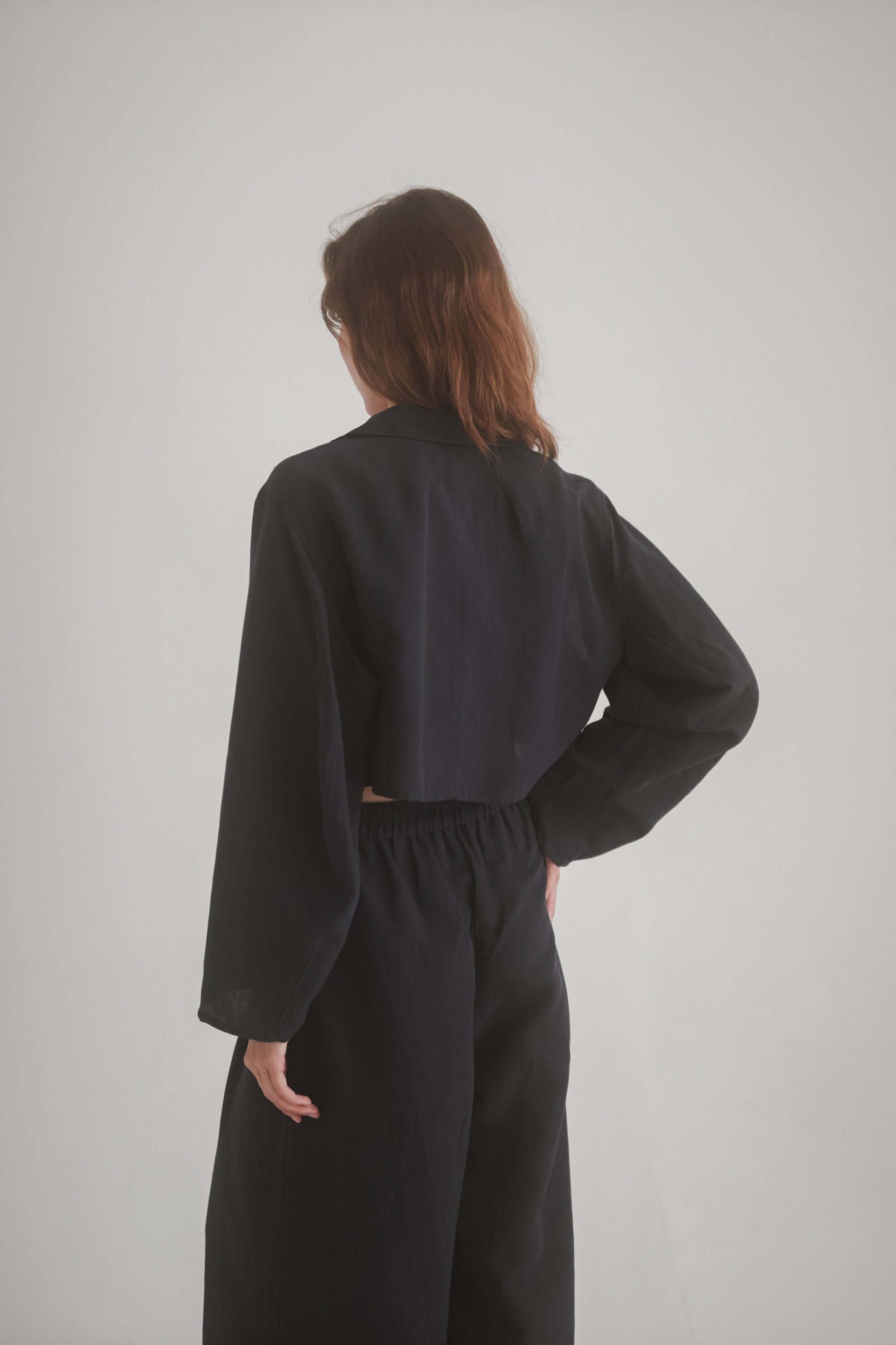Back view of a woman wearing a black cropped jacket with wide trousers, showcasing the stylish fit and armhole cutouts.