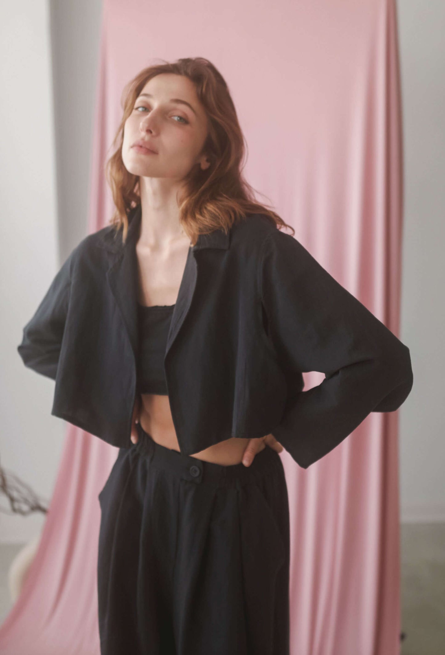 Model wearing a black cropped jacket styled with wide trousers against a pink backdrop.