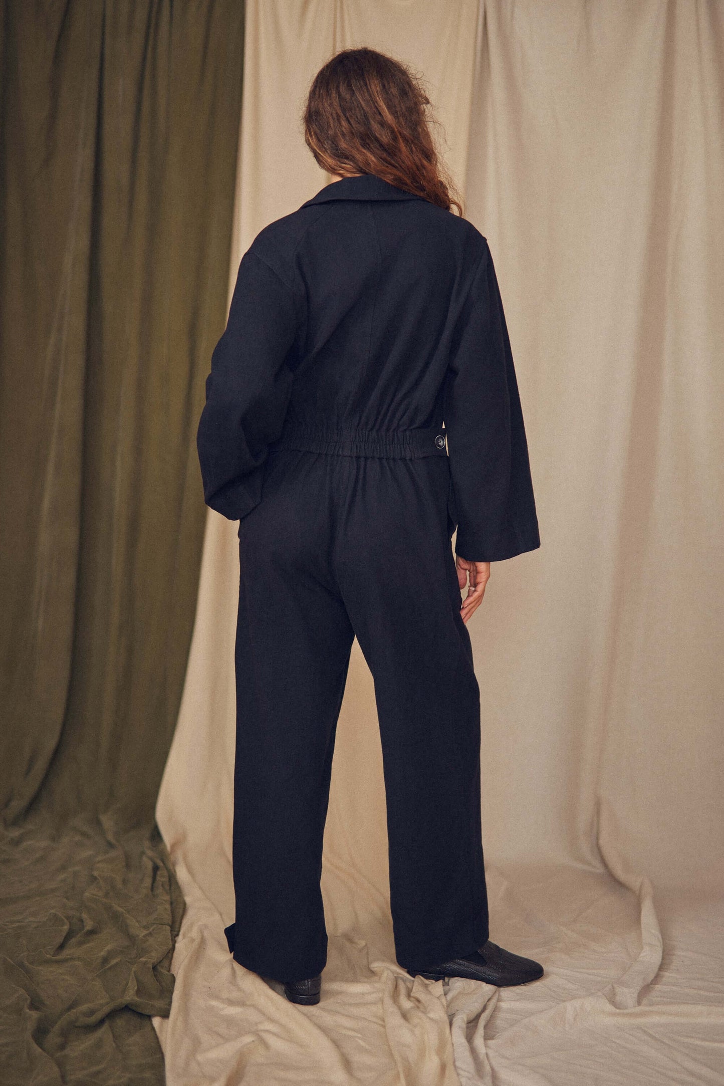 Person wearing a black jumpsuit standing against a beige curtain. Person wearing a black jumpsuit standing against a beige curtain.