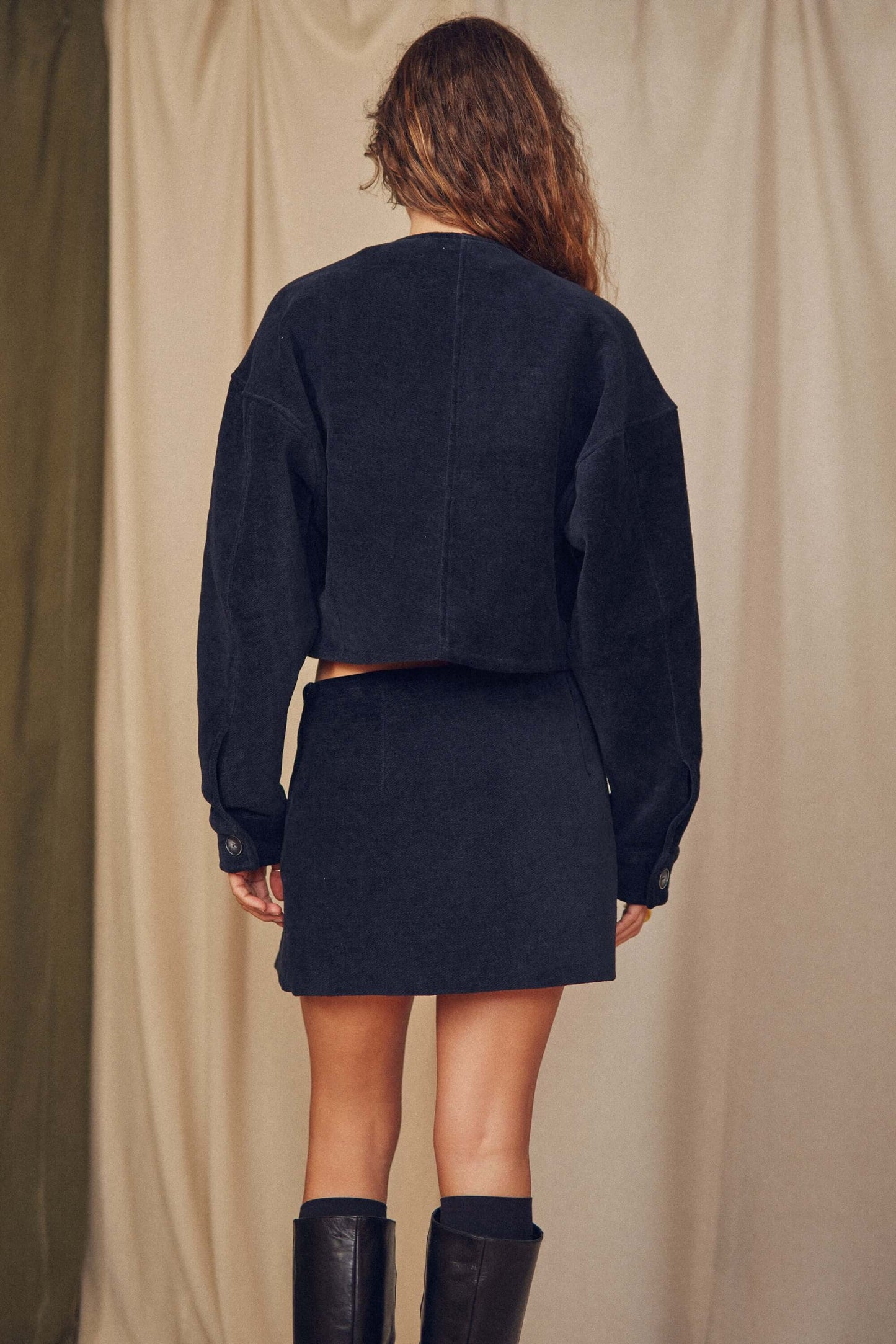 Back view of a model wearing a black organic cotton crop jacket and matching skirt, showcasing a bomber-inspired style.