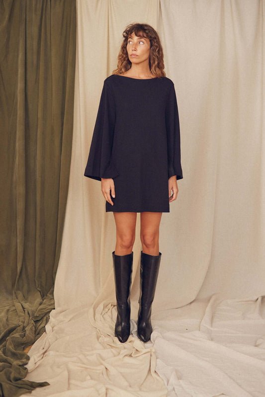 Model wearing a black organic cotton mini dress with flared sleeves, styled with tall black boots.