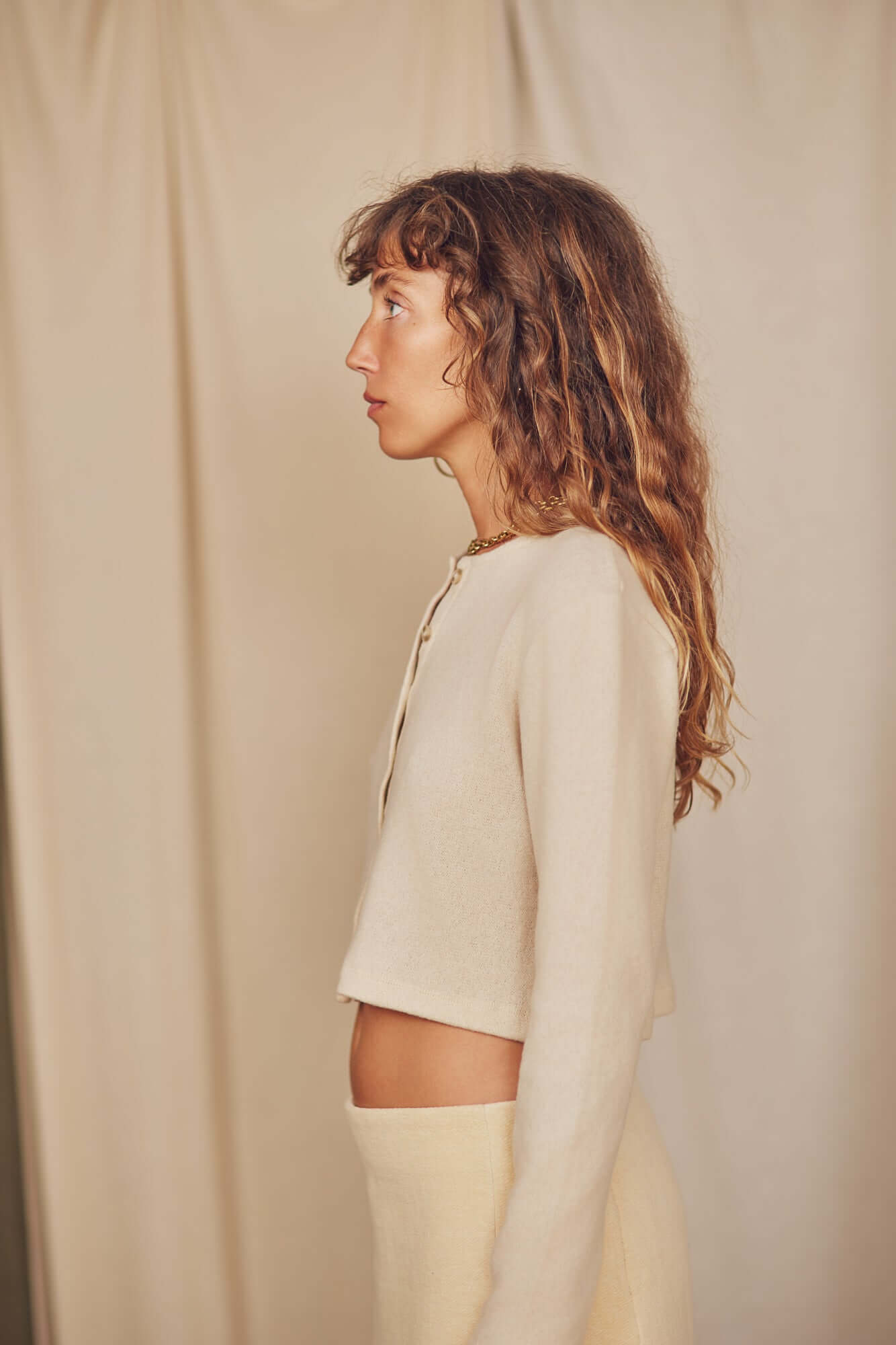 Profile view of a model wearing an Oat Wool Crop Knit Cardigan, showcasing its cropped length and soft oat color.