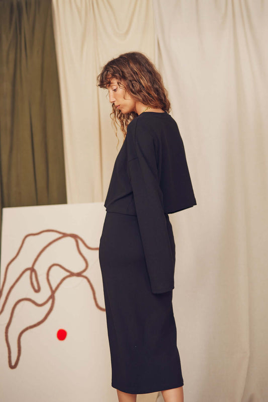 Model wearing a black organic cotton midi skirt paired with a long-sleeve top in a minimalist setting.