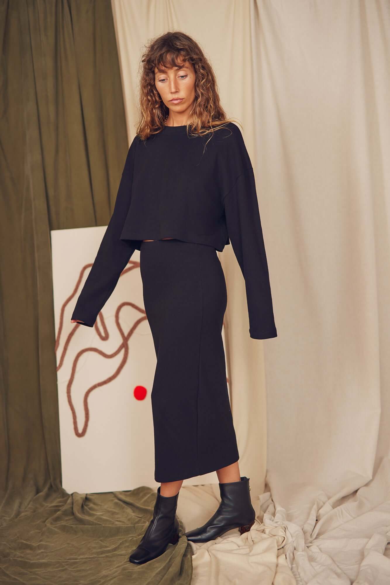 Model wearing a black organic cotton midi pencil skirt with a matching top, styled for autumn/winter fashion.