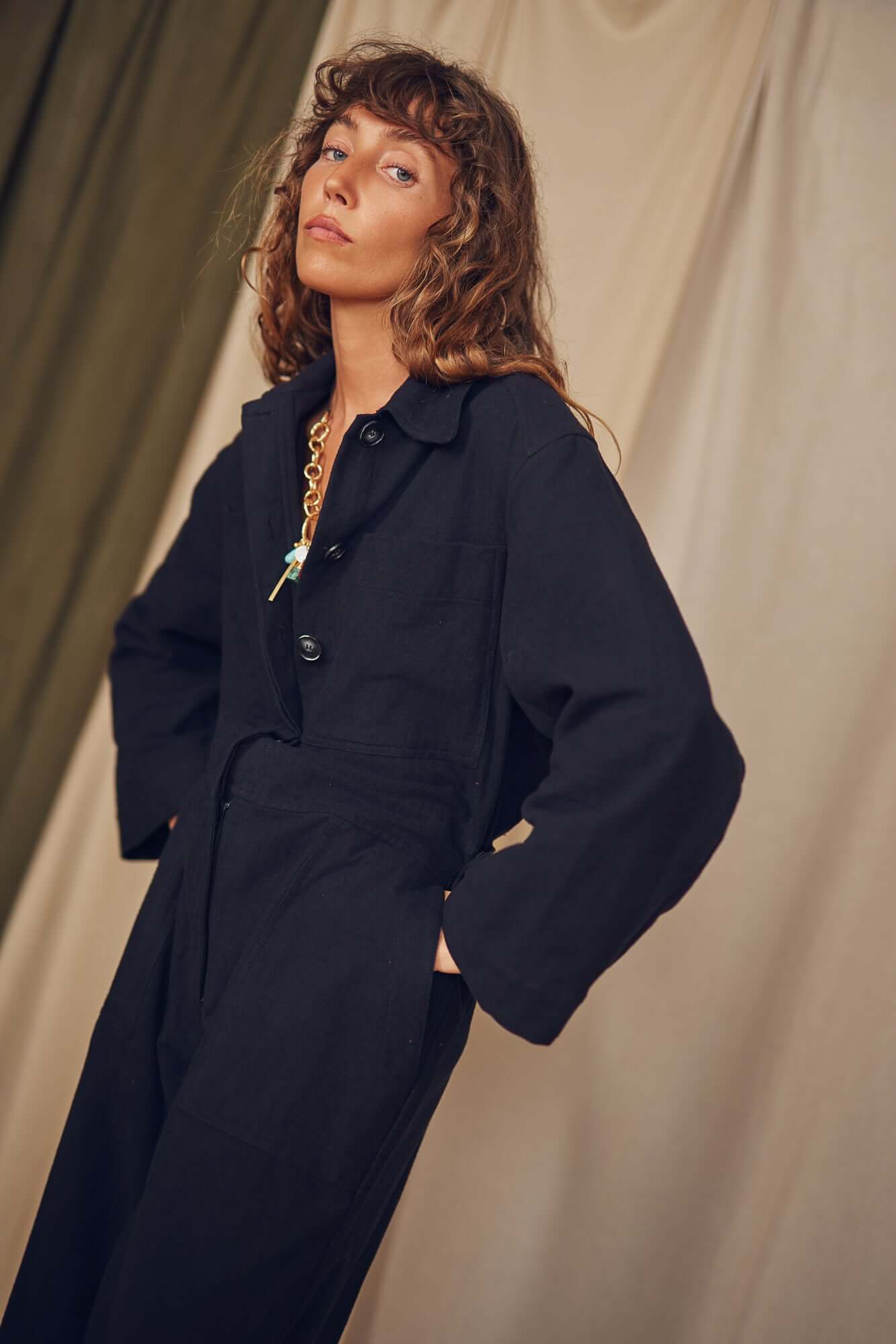 Person wearing a black jumpsuit standing against a beige curtain. Person wearing a black jumpsuit standing against a beige curtain.
