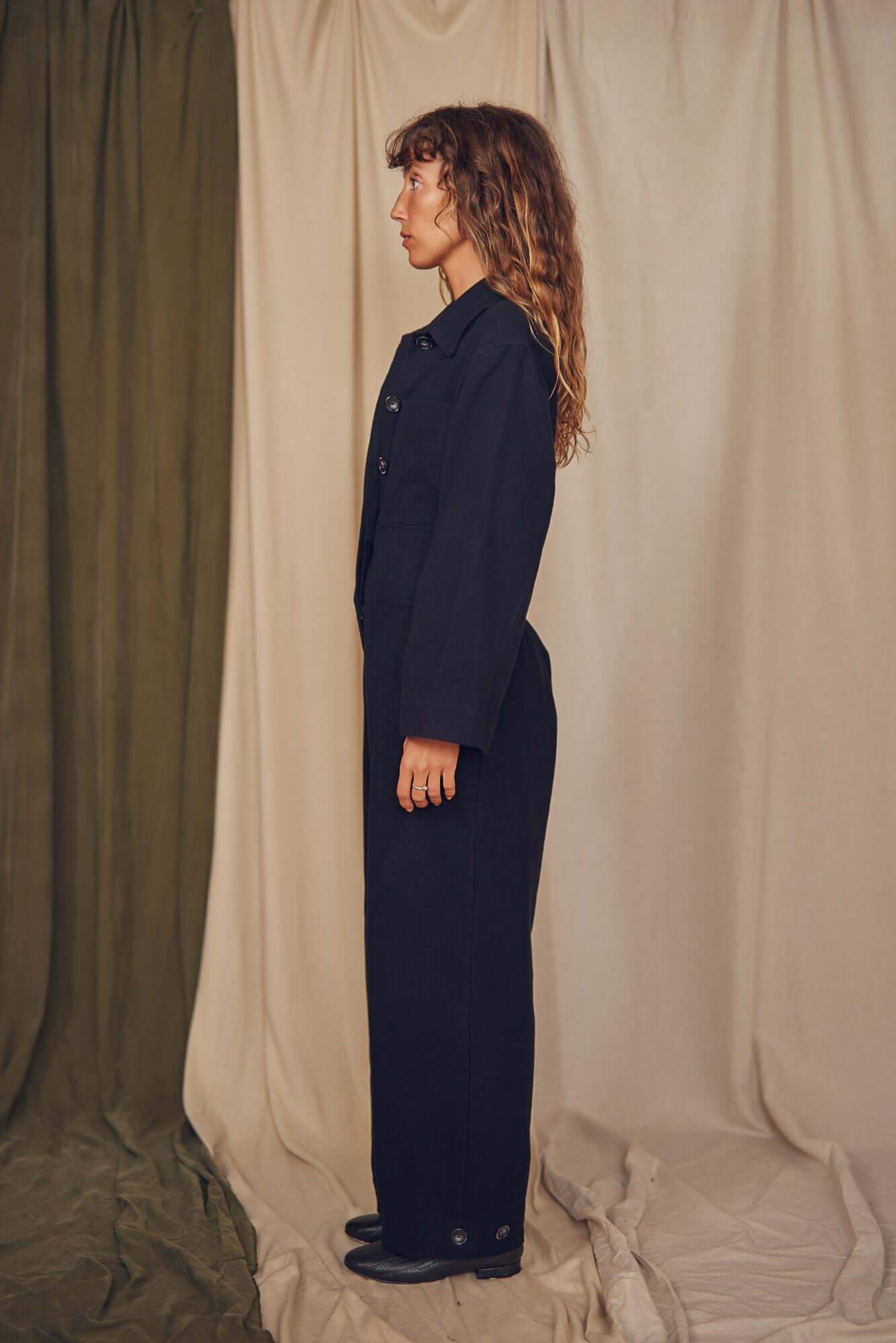 Person wearing a black jumpsuit standing against a beige curtain. Person wearing a black jumpsuit standing against a beige curtain.