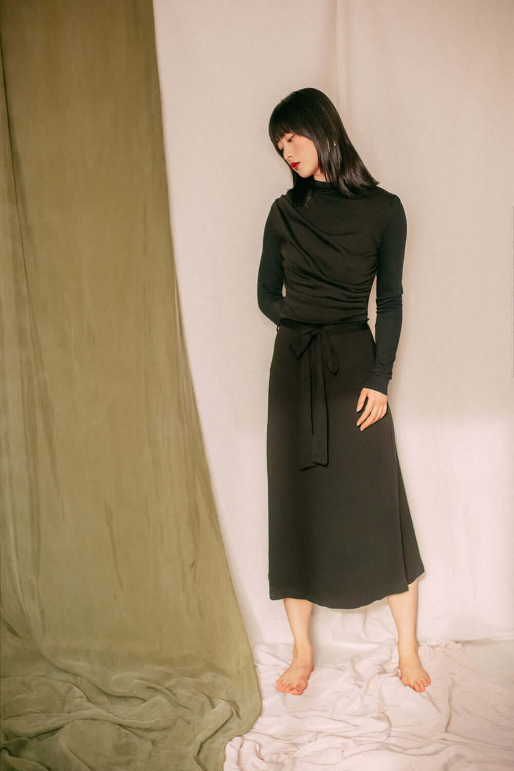 Woman wearing black drape knit dress, online exclusive, fine knit material, midi length with drape bust detail, standing barefoot indoors