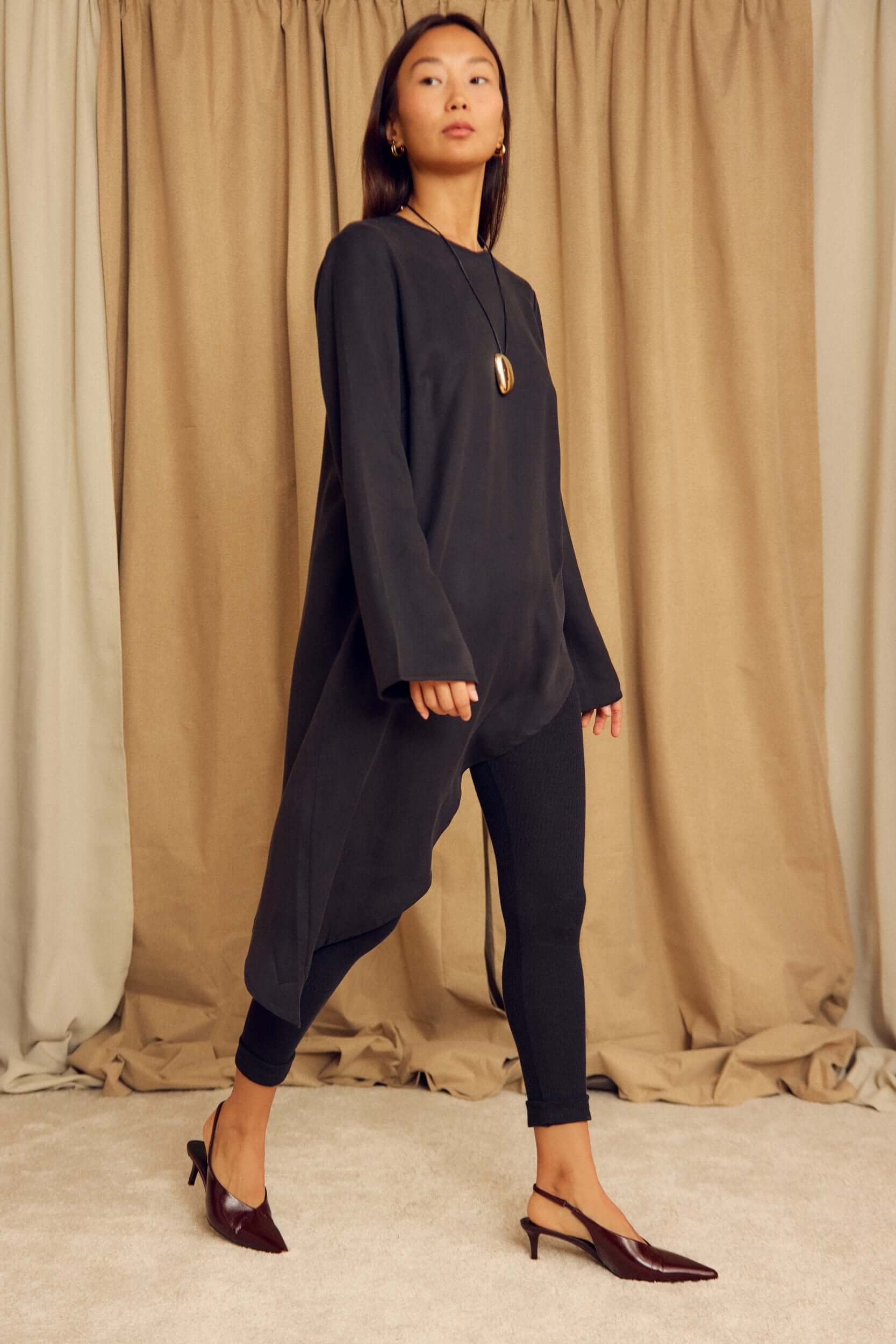 Woman wearing a Black Lyocell Asymmetric Tunic paired with leggings and heels, showcasing its unique silhouette.