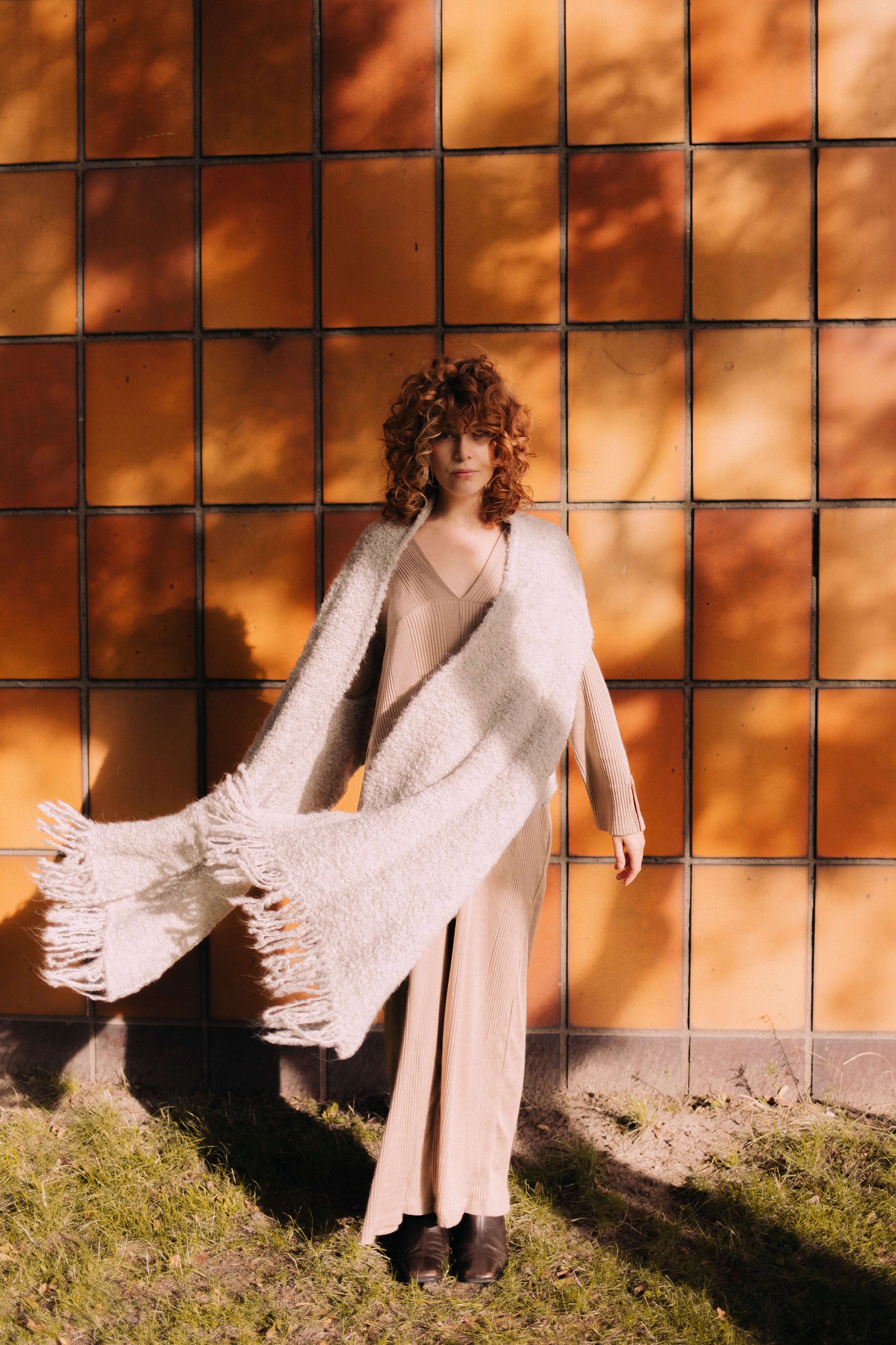 Woman wearing GREY PONCHO SCARF styled as scarf in sunlight, showcasing soft texture and eco-friendly design against a tiled wall background.