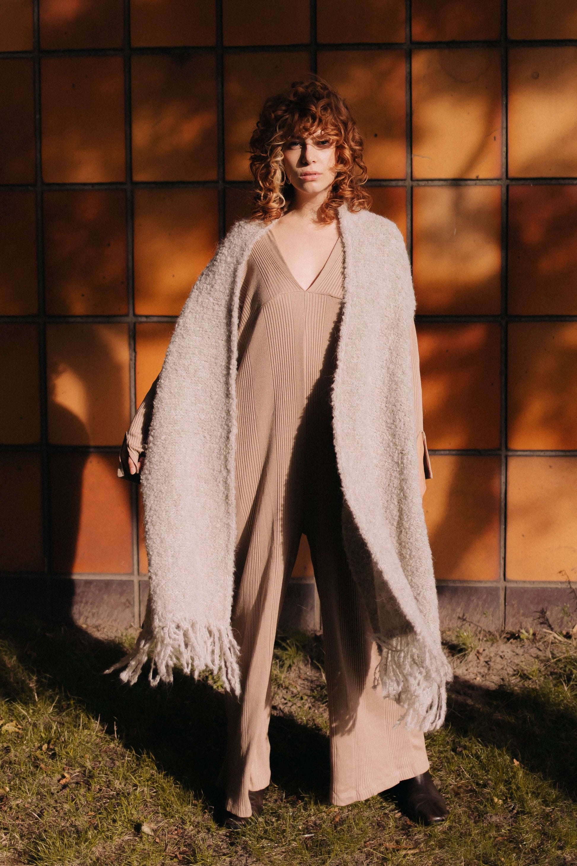 Woman wearing grey poncho scarf made of sustainable alpaca and recycled polyester yarn, stylishly draped over a beige jumpsuit.