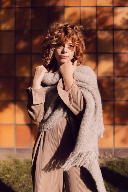 Woman wearing a grey poncho scarf made from sustainable alpaca and recycled polyester, standing against a sunny tiled wall.