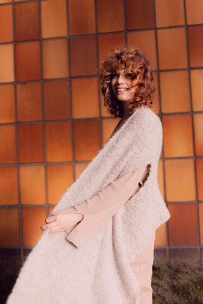 Woman wearing grey poncho scarf made from sustainable alpaca and recycled polyester, standing against an orange tiled wall.