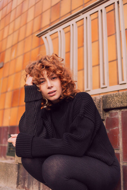 Woman wearing a cozy black sweater with a high neck, sitting against an orange tiled wall.