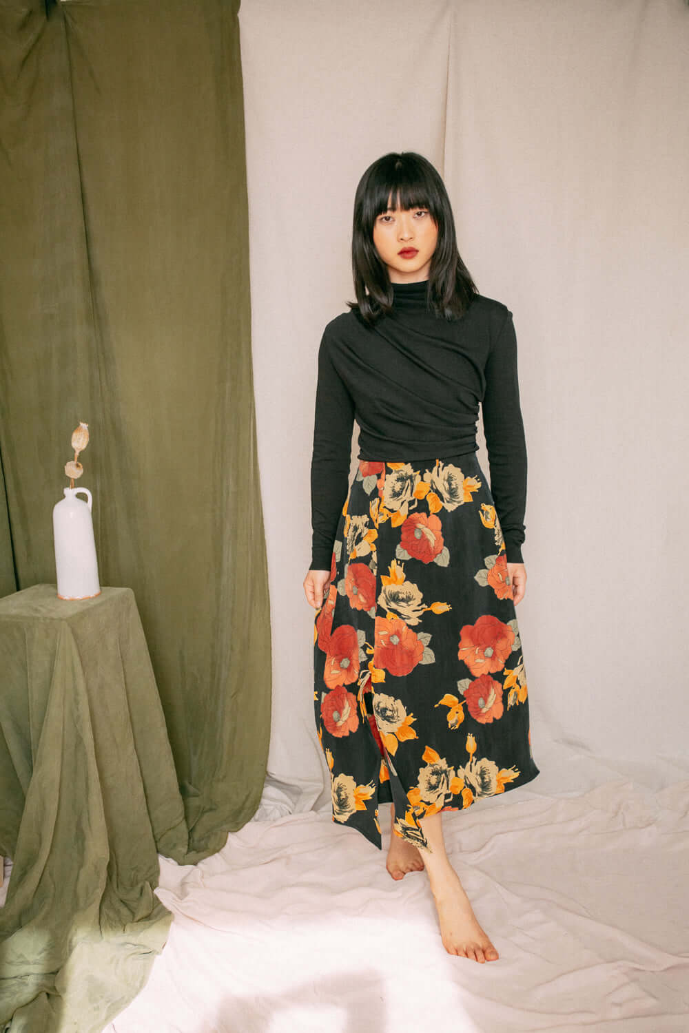 Woman wearing black drape knit top with floral skirt, standing in a cozy room with green and white fabric background.