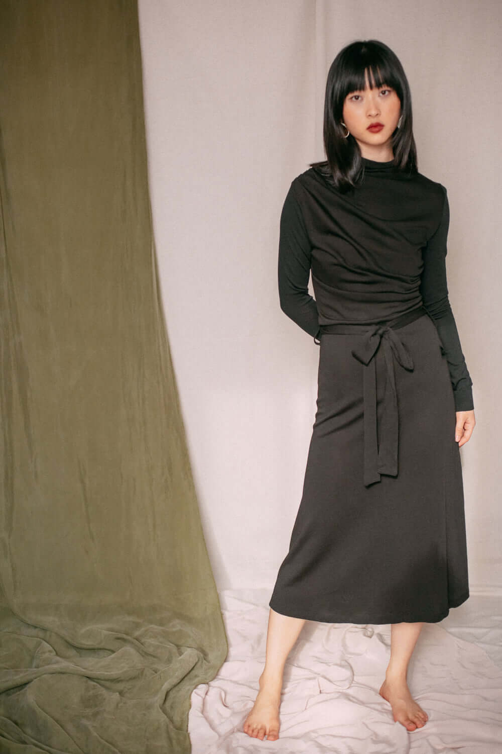 Woman wearing a black drape knit dress with a midi length and draped bust detail, made from lyocell and organic cotton jersey.