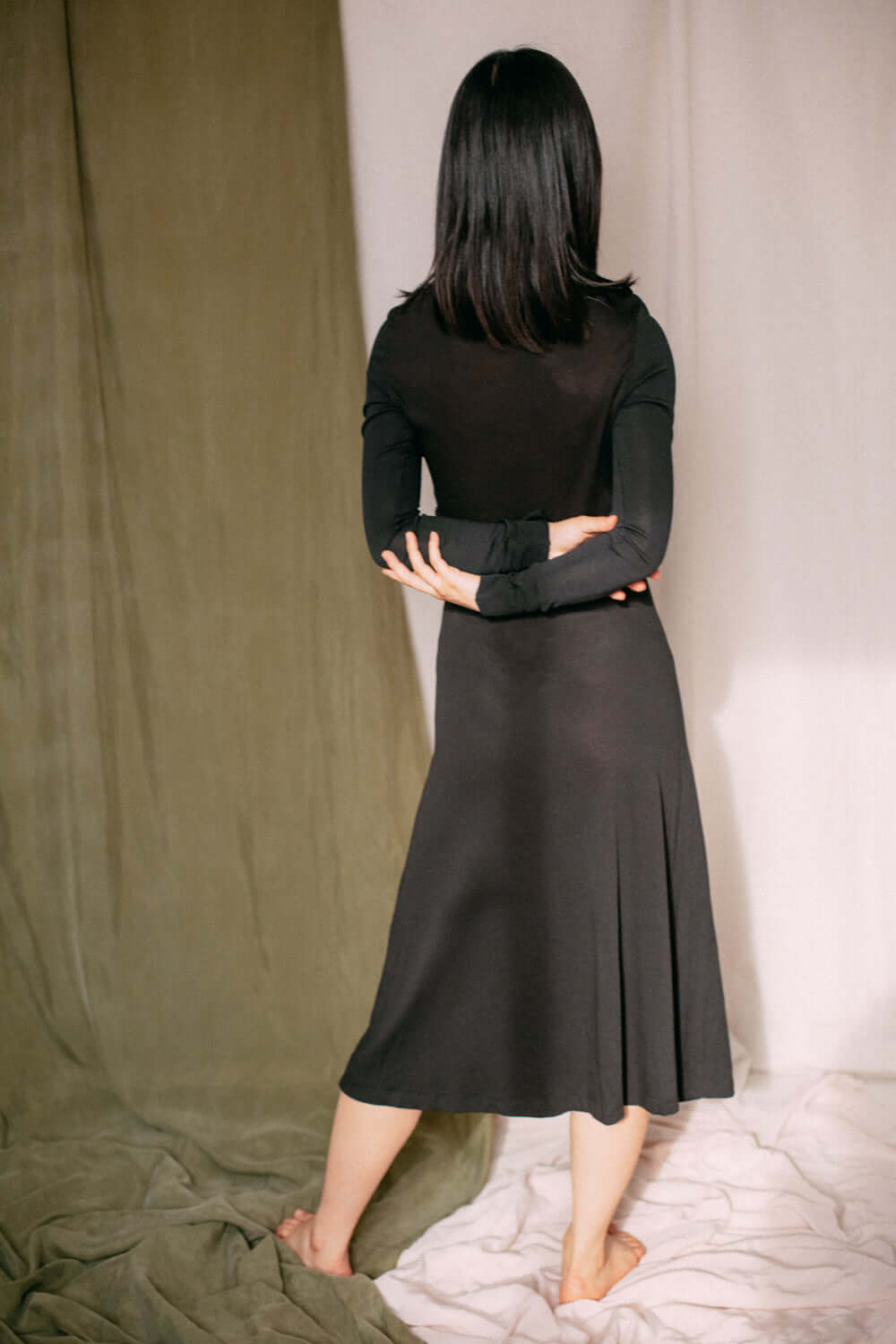 Rear view of woman wearing black drape knit dress with midi length, long sleeves, and draped bust detail.