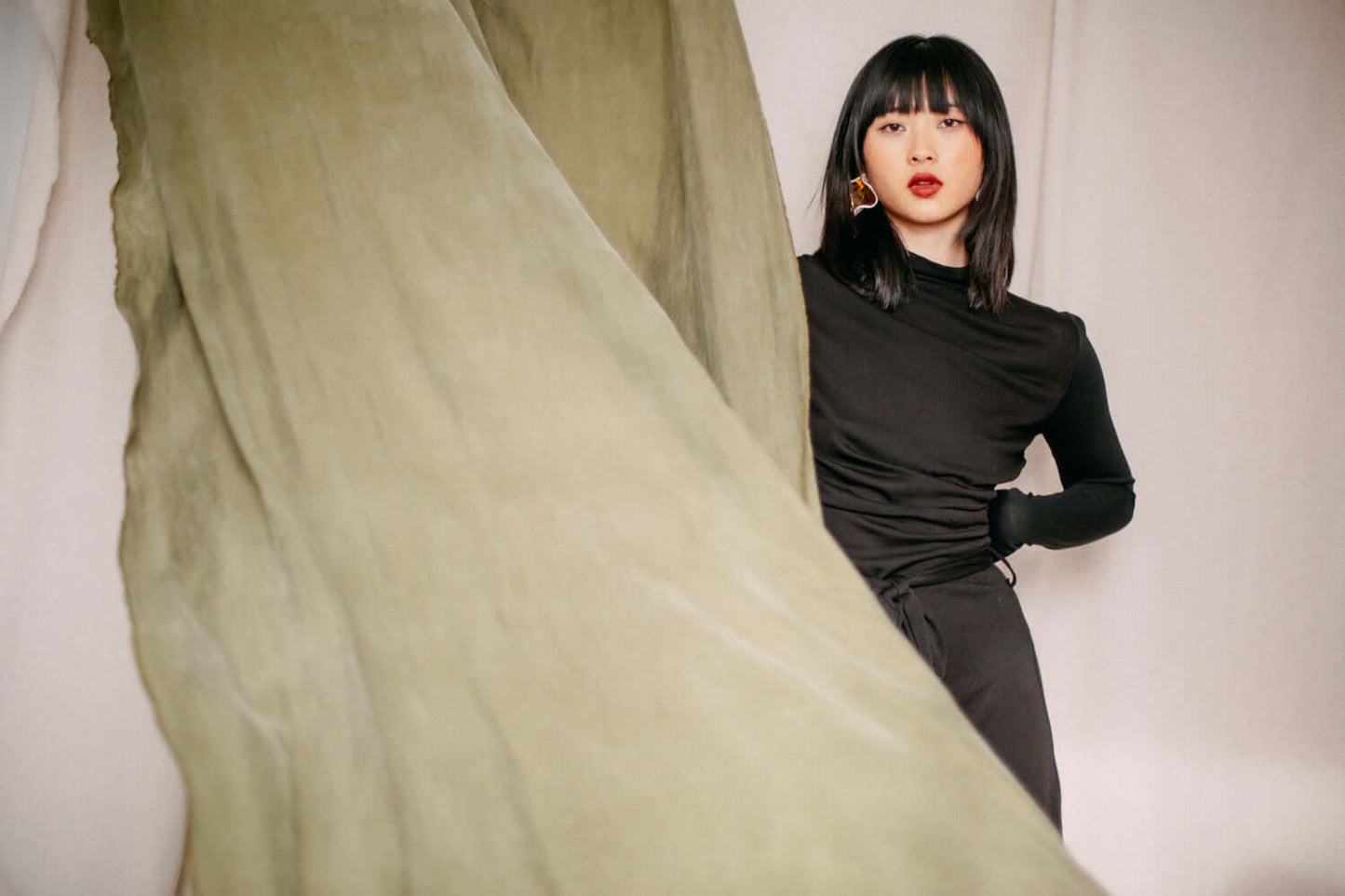 Woman wearing black drape knit dress with feminine bust detail, standing behind flowing green fabric curtain