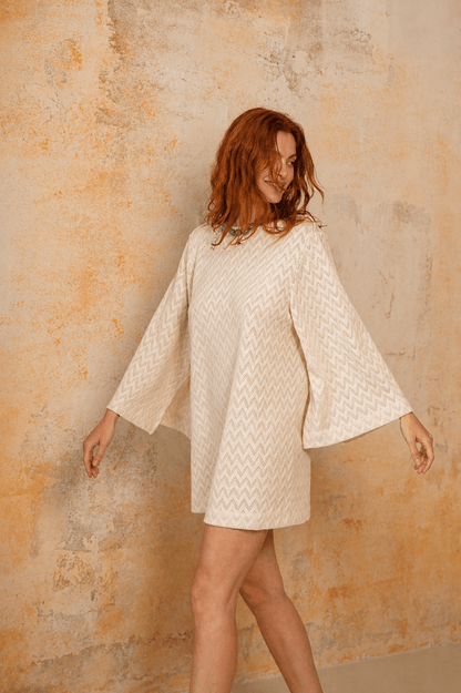 Model wearing Organic Cotton Crochet Mini Dress with flared sleeves in ecru against a textured background.