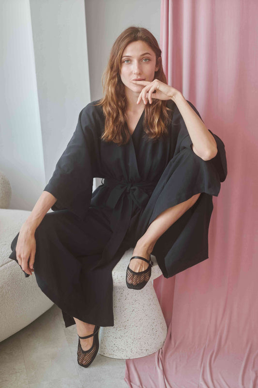 Model wearing a black kimono jumpsuit with a belt, sitting on a sculptural chair against a soft pink backdrop.