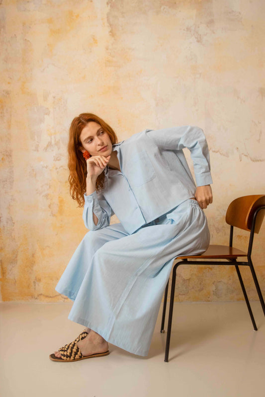 Organic Cotton Wide Culottes in Pale Blue worn by a model sitting on a chair against a textured wall.