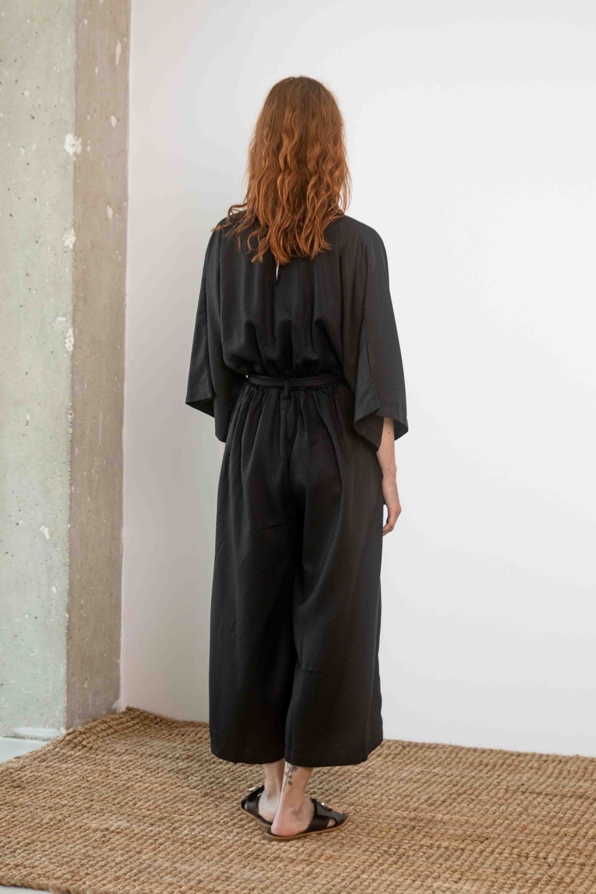 Back view of a model wearing the Tencel Kimono Jumpsuit in Black, showcasing its relaxed fit and culotte-style trousers.