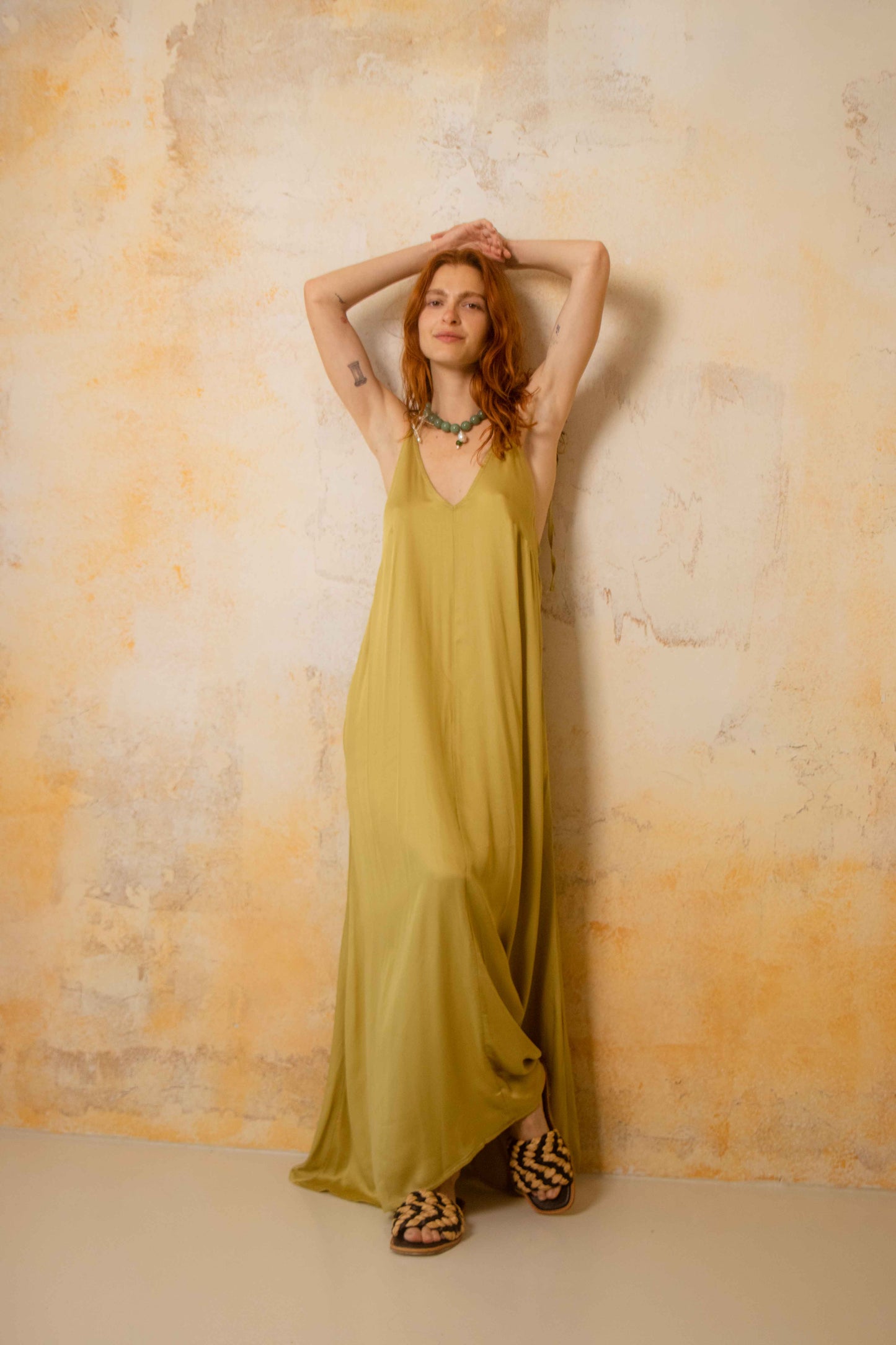 Model wears a Ecovero Maxi Dress, showcasing a lightweight, olive-colored design on a textured background.