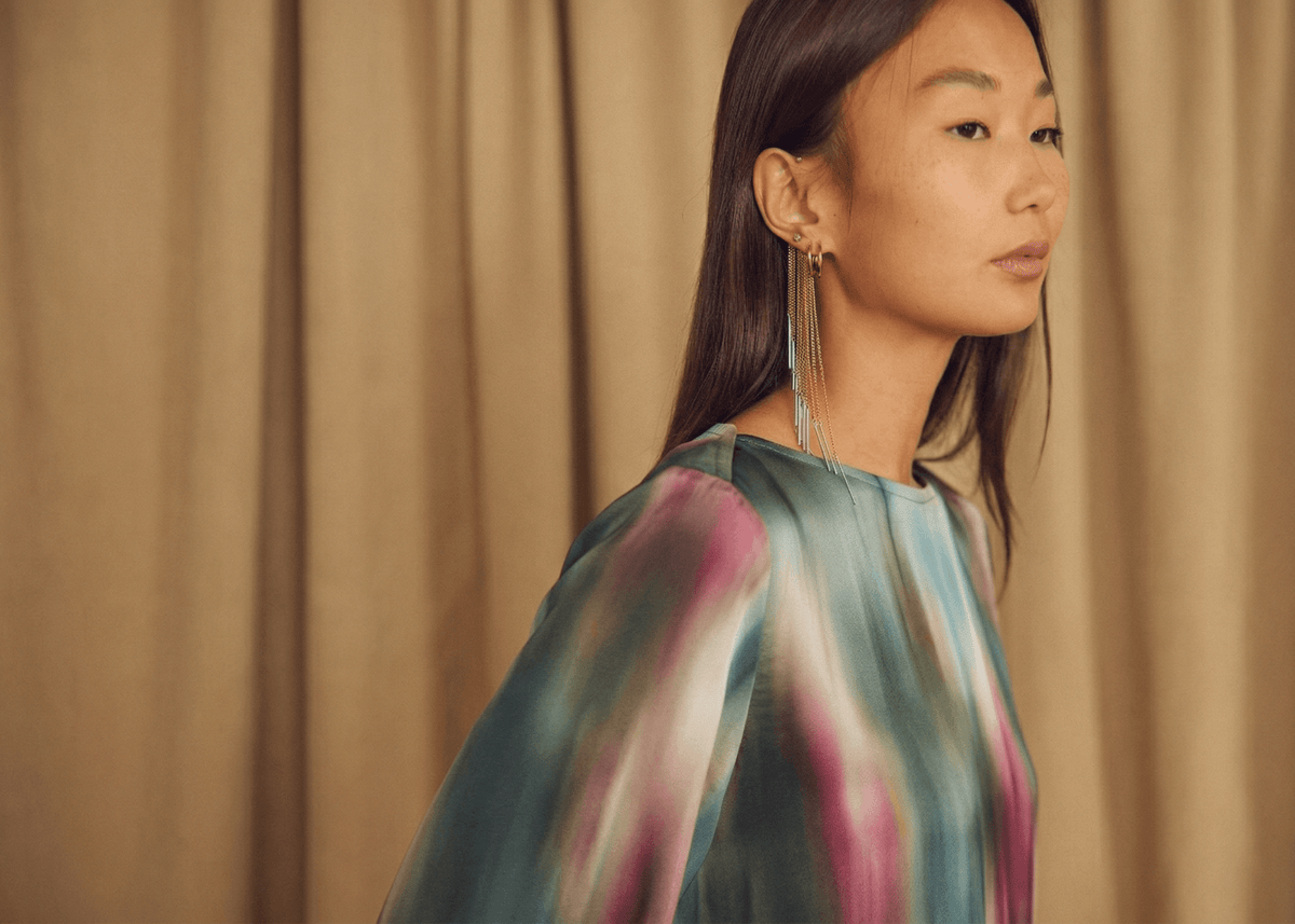 Close-up of a woman in a colorful blouse with long earrings, posed against a soft brown backdrop.