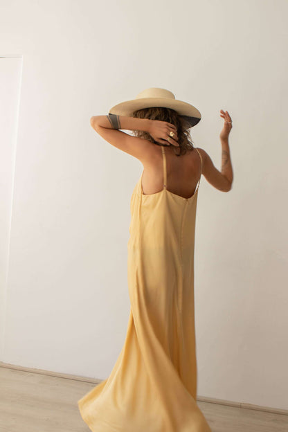 Model wearing a vanilla maxi dress with adjustable straps and sun hat, showcasing flowy design from the back.
