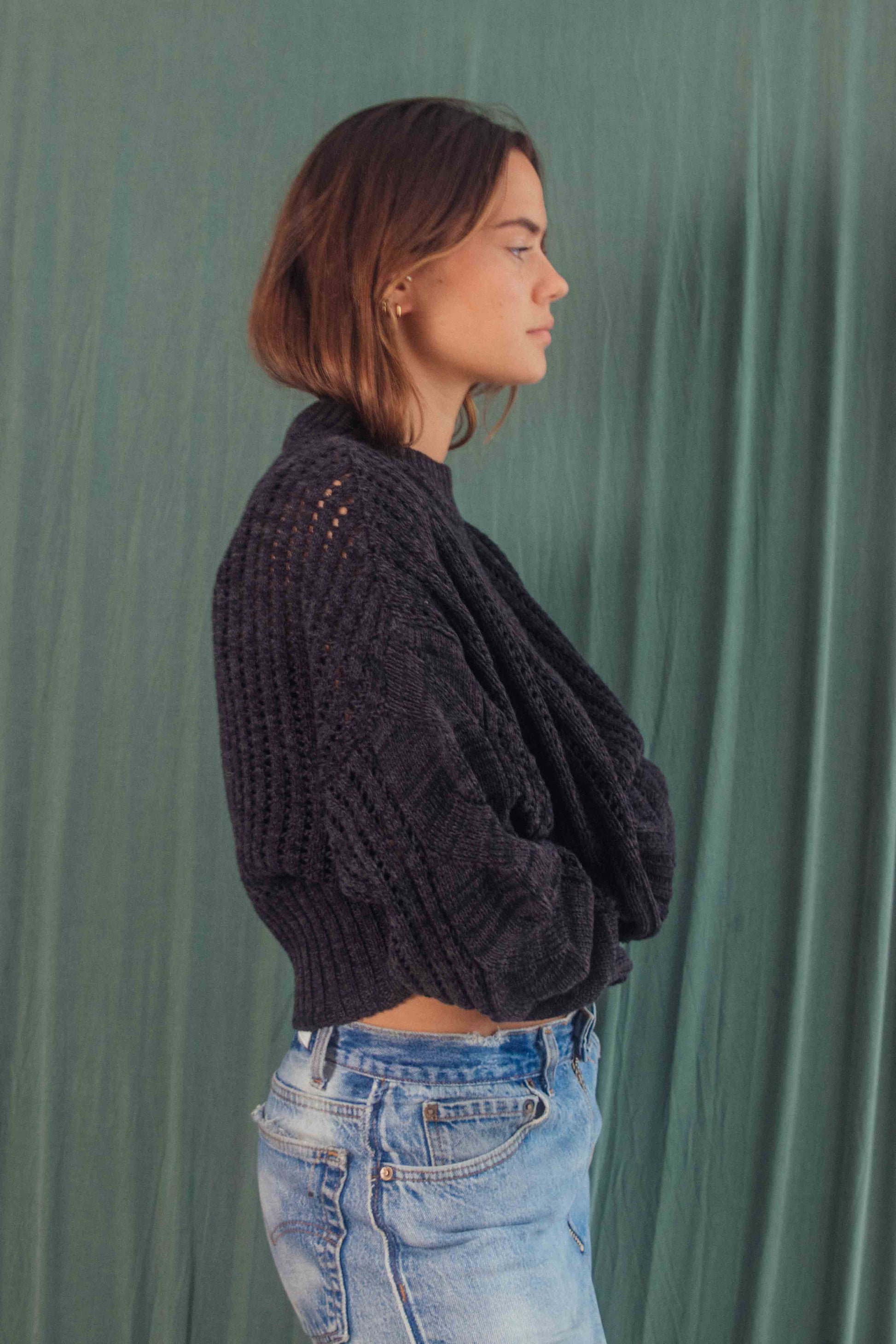 Woman wearing melange black sweater made from 100% organic cotton, crop & wide fit, standing against a green background.