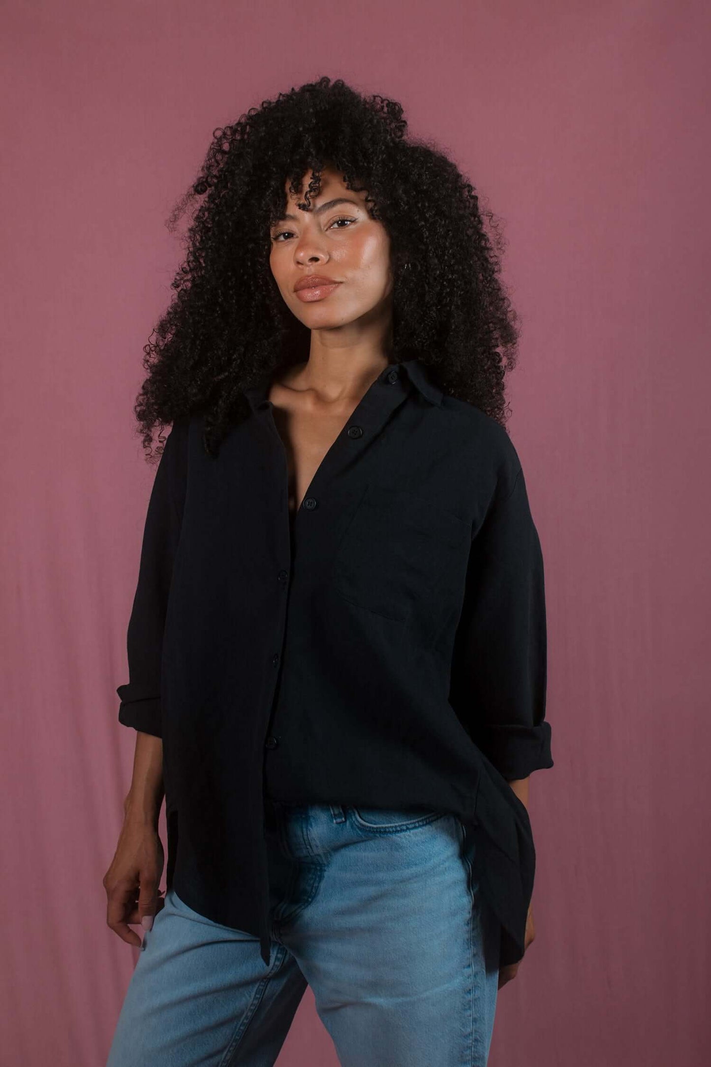 Model wearing a black wide shirt made from organic cotton, styled with jeans against a pink background.