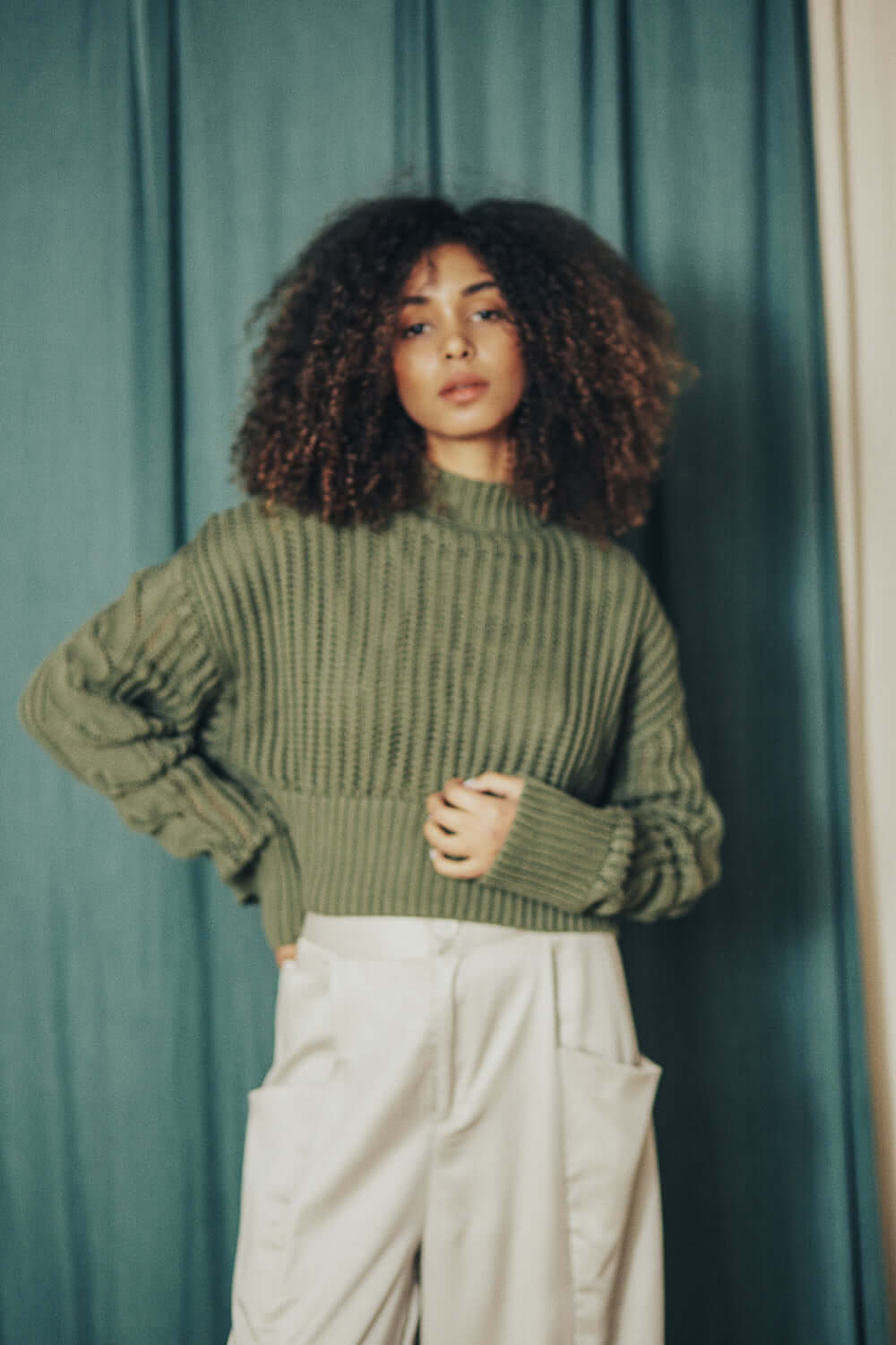 Woman wearing sage green sweater made from 100% organic cotton yarn, standing against a green curtain. Crop and wide fit.