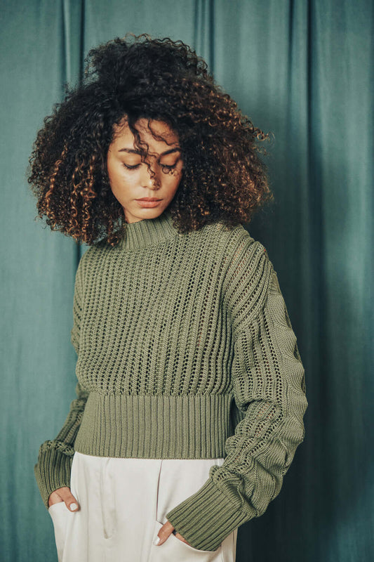 Woman wearing sage green cropped sweater made from 100% organic cotton yarn, wide fit, styled with white trousers against a green curtain backdrop.