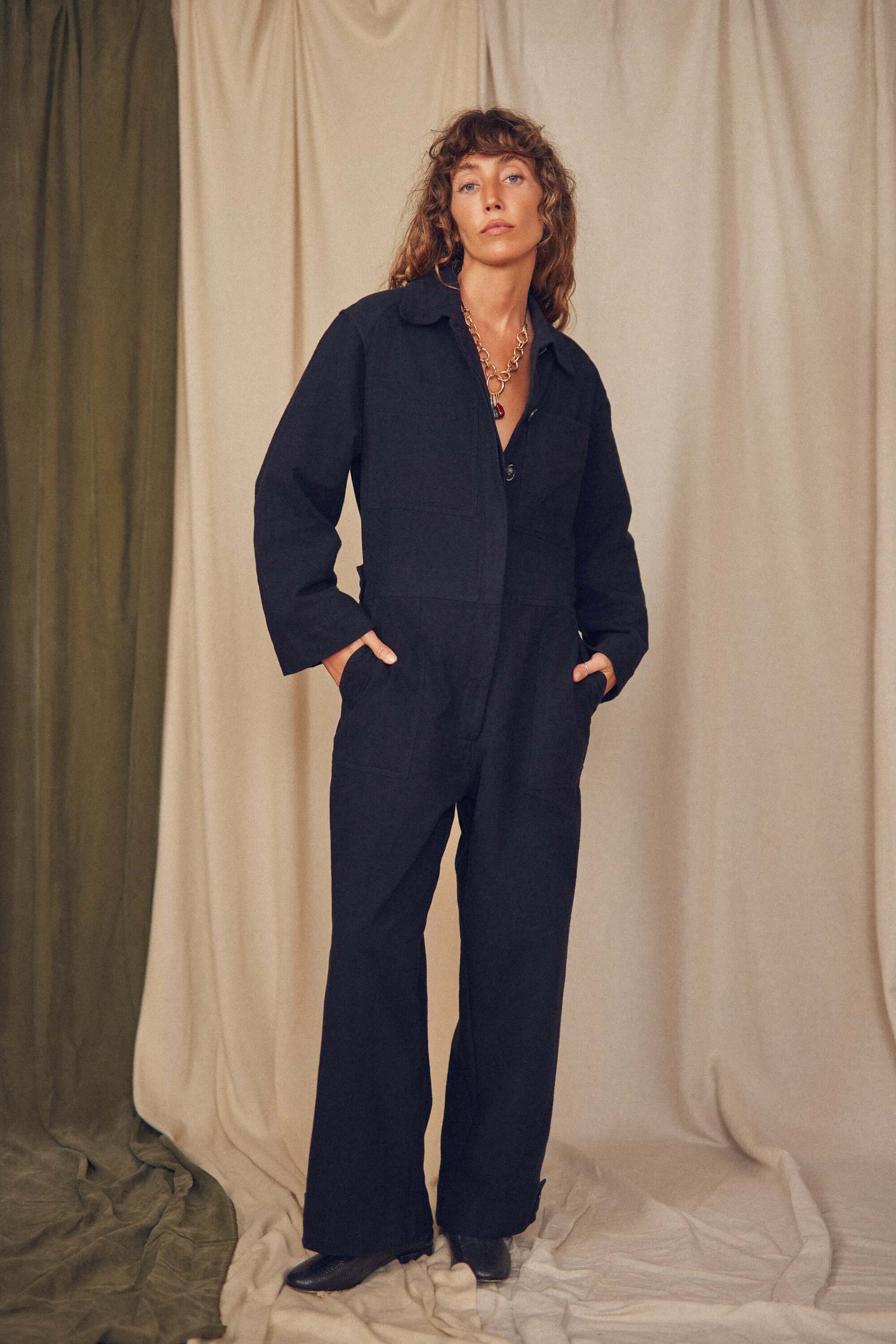 Person wearing a black jumpsuit standing against a beige curtain.