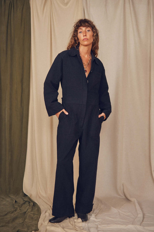 Person wearing a black jumpsuit standing against a beige curtain.
