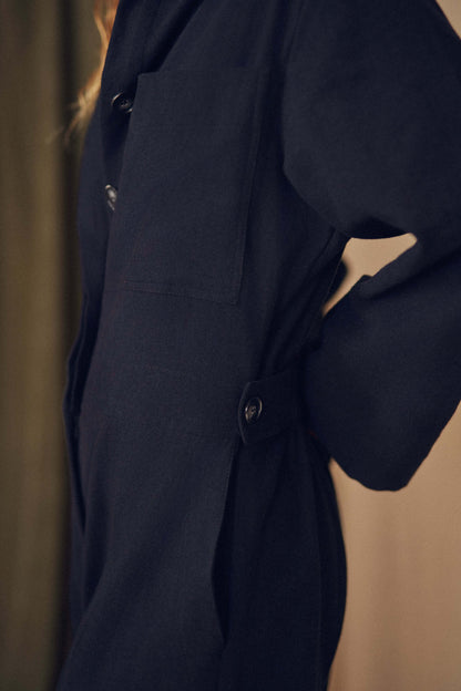 Person wearing a black jumpsuit standing against a beige curtain. Person wearing a black jumpsuit standing against a beige curtain.