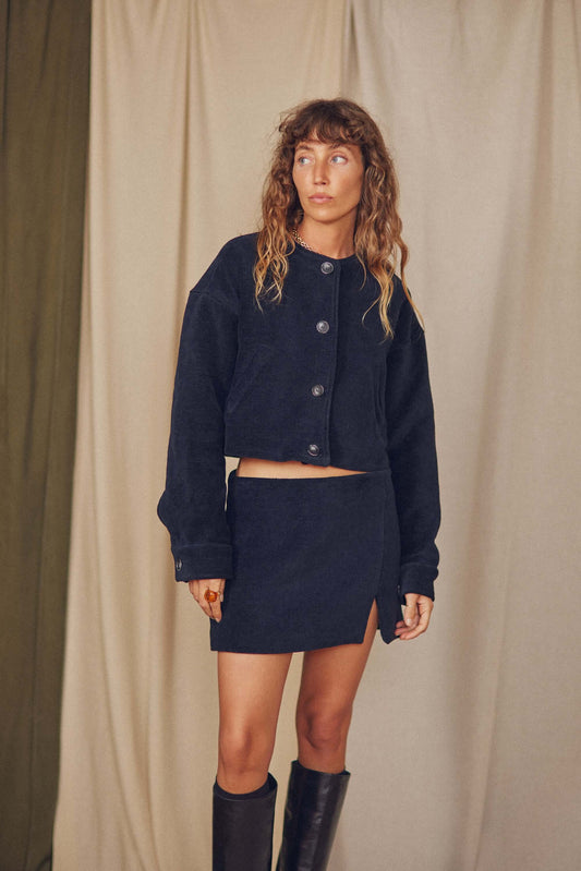 Model wearing a black organic cotton mini skirt and matching cropped jacket, featuring a thigh split for a stylish autumn look.