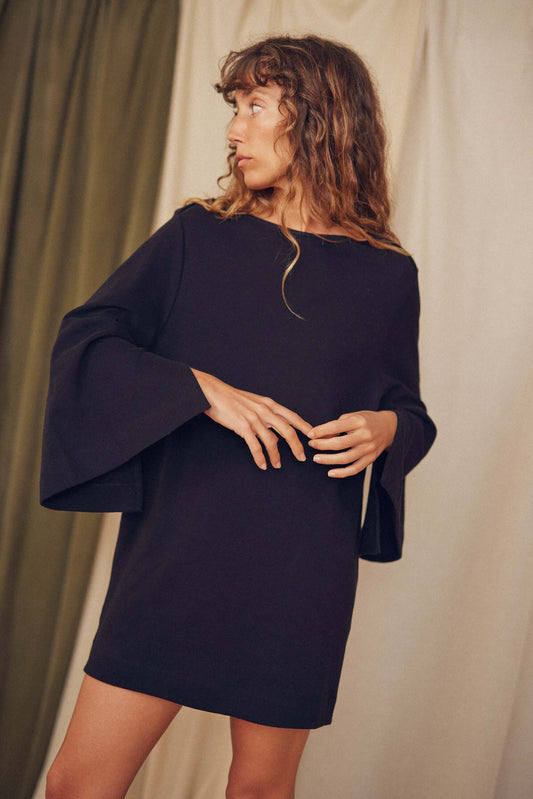 Model wearing a black organic cotton mini dress with flared sleeves against a textured backdrop.