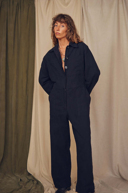Person wearing a black jumpsuit standing against a beige curtain. Person wearing a black jumpsuit standing against a beige curtain.