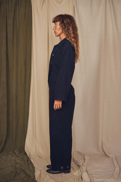 Person wearing a black jumpsuit standing against a beige curtain. Person wearing a black jumpsuit standing against a beige curtain.