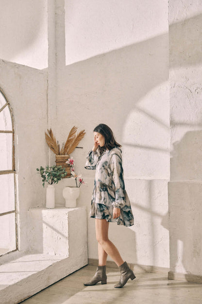 Woman wearing a flowy printed mini dress standing by a sunlit window in neutral-toned room.