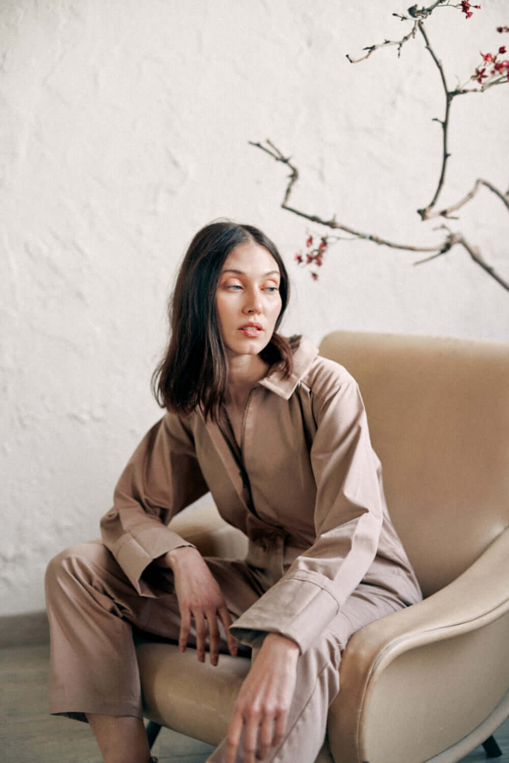 Woman wearing taupe denim boilersuit made of organic cotton, sitting on a chair, with turned cuffs and a minimalist background.