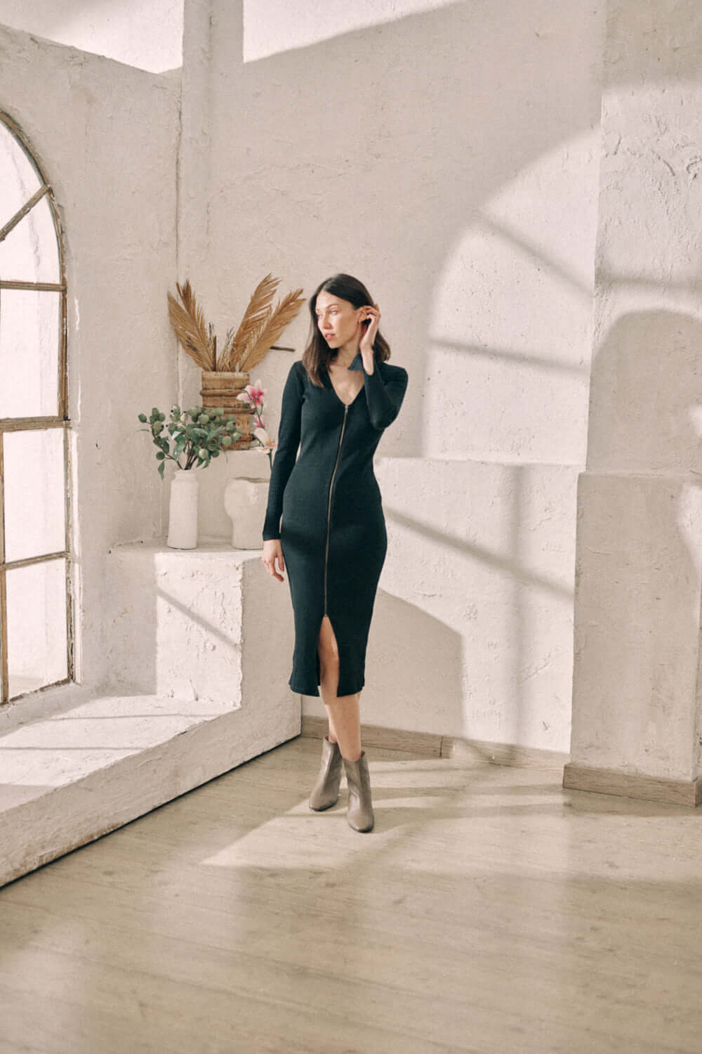 Woman wearing black reversible zip dress made from sustainable jersey, standing in a sunlit room with minimalist decor.