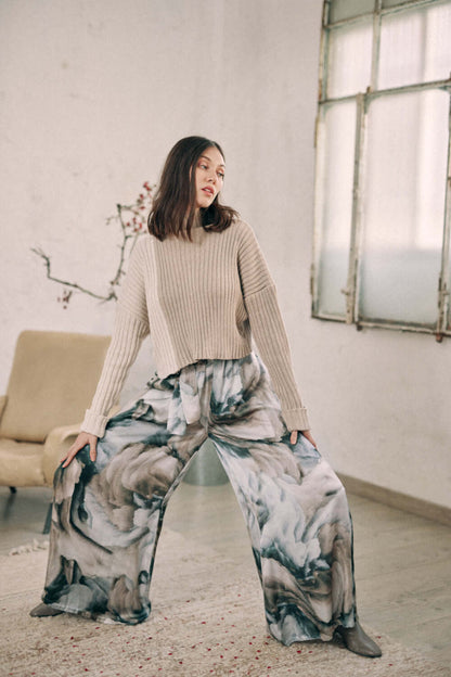 Woman wearing a bicolor beige sweater with high neck, styled with artistic wide-leg pants, standing in a minimalist room.