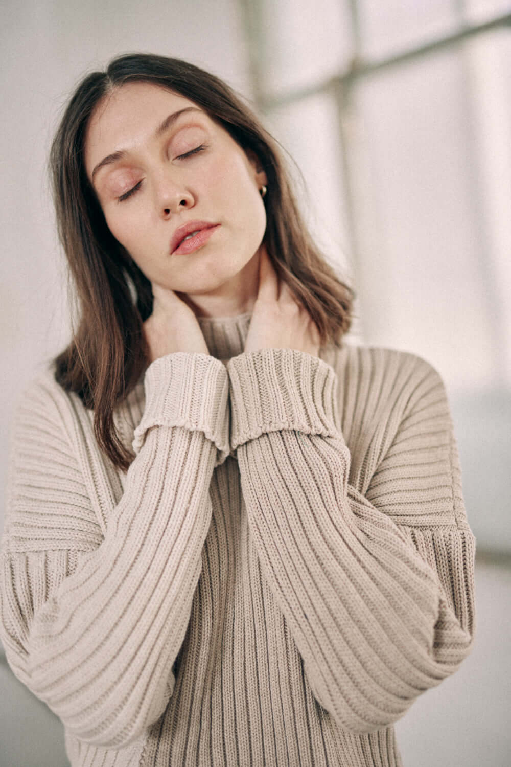 Woman wearing a bicolor beige sweater made of organic cotton, featuring a high neck and wide fit design, in a cozy setting.