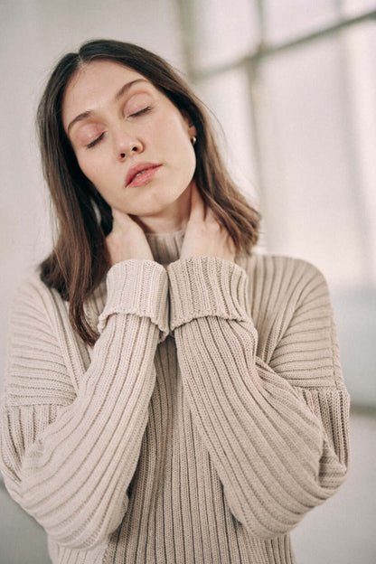 Woman wearing a bicolor beige sweater made of organic cotton, featuring a high neck and wide fit design, in a cozy setting.