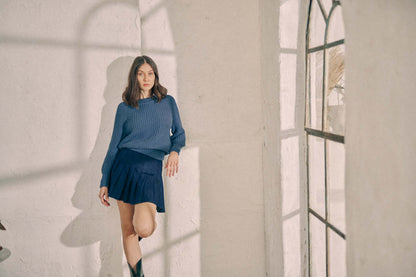 Model wearing navy sweater with skirt, standing by a sunlit window, showcasing online exclusive vegan organic cotton fashion.